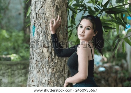 A stylish young woman dressed in goth inspired clothing poses gracefully by a tree in a serene forest setting, exuding a mysterious and enchanting vibe.