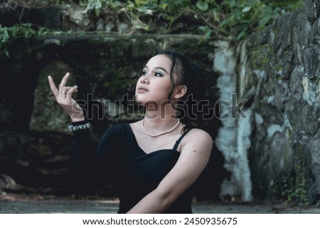 A young woman in goth-inspired attire stands in a forest, her expression introspective as she gracefully poses among the rustic ruins.