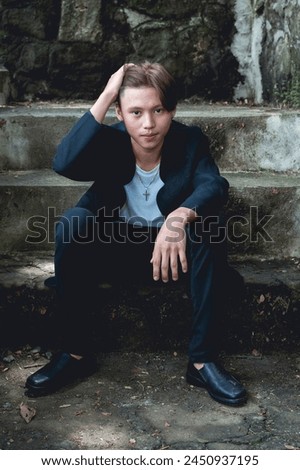 An Asian male in his youth wearing goth-style clothing, posed against some old stairs outdoors, exuding a mysterious aura.