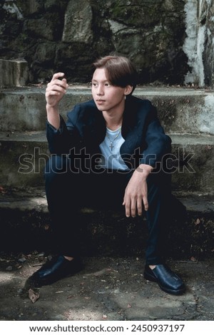 A young Asian man dressed in stylish goth-inspired clothing sits on steps in a forest, looking thoughtfully at something in his hands.