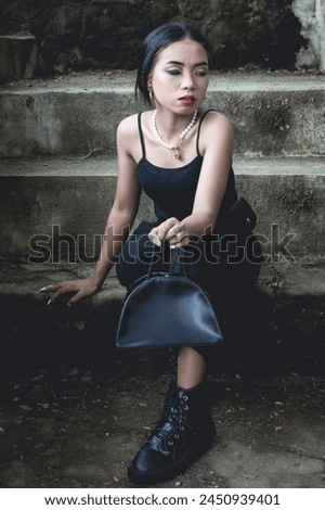 A young woman dressed in goth-inspired fashion sits on concrete steps in a forest, exuding a moody and mysterious vibe.