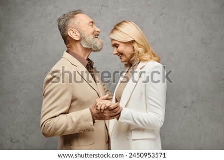 Mature, elegant couple in debonair attire embrace, holding hands lovingly against a gray backdrop. Royalty-Free Stock Photo #2450953571