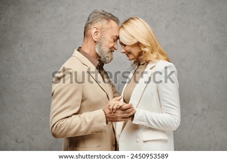 Mature man and woman, elegant in debonair attire, stand side by side in a loving pose on a gray backdrop. Royalty-Free Stock Photo #2450953589