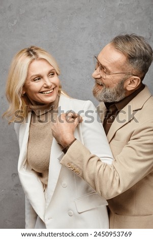 A elegant, mature man helps a woman put on her coat in a debonair pose against a gray backdrop. Royalty-Free Stock Photo #2450953769
