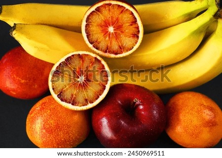 Several ripe yellow bananas next to a red apple, oranges on a dark background