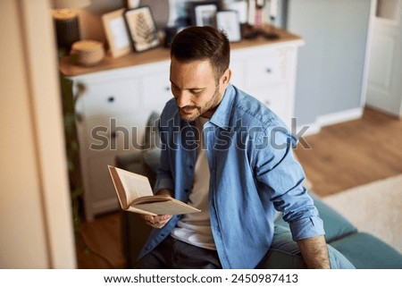 A top view of a young adult man taking a break and reading a book novel in his living room while leaning on a couch.