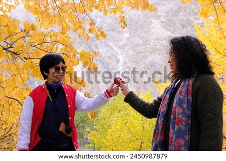 Two Asian female sisters holding an apple, posing for a picture with a view of the colorful foliage in the morning light in Passu Valley, Gilgit-Baltistan, Pakistan.