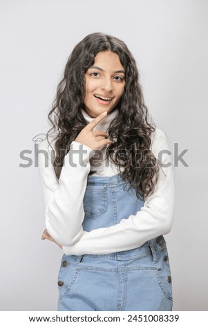 Closeup portrait of young Pakistani girl with wavy loose hair pictured isolated on white background dressed in denim jumpsuit, smiling happily at camera feeling joy and satisfaction