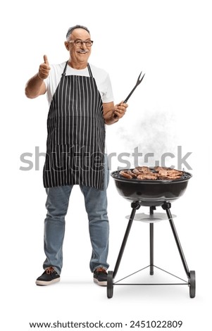 Mature man next to a portable barbecue grill holding a fork and gesturing thumbs up isolated on white background