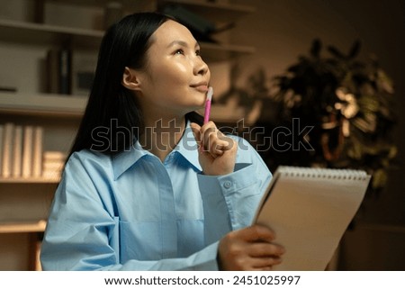 A young Asian woman is engrossed in deep thought as she holds a pen and notepad, looking for inspiration in a cozy home library filled with books. Soft lighting enhances the contemplative mood as the