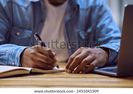 Man, hands and writing with laptop in book for studying, elearning or education on desk at home. Closeup of male person, student or writer taking notes in diary for reminder, information or agenda Royalty-Free Stock Photo #2451041509