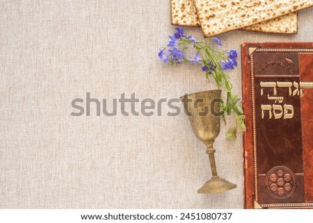 Jewish holiday Pesach (Passover) celebration concept. Traditional book with text in Hebrew: "Passover Haggada" ("Passover Tale"), Kiddush cup, matzah on cotton fabric background. Royalty-Free Stock Photo #2451080737