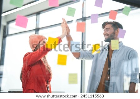 Business team uses a glass wall covered with sticky notes to outline processes during meeting. Engaged colleagues gather around a glass brainstorming board. plotting ideas and strategies. Royalty-Free Stock Photo #2451115563