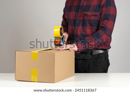 Storekeeper seals box with duct tape. Cropped warehouse employee with package. Storekeeper uses dispenser with adhesive tape. Guy in checkered shirt packs goods. Storekeeper prepares box for delivery Royalty-Free Stock Photo #2451118367