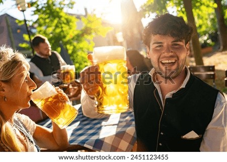 Joyful young man toasting with beer mug at Oktoberfest or beer garden, dressed in traditional attire or tracht in German, bright sunlight in background Royalty-Free Stock Photo #2451123455
