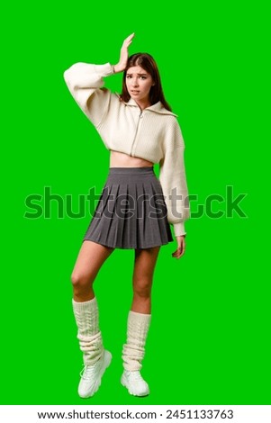A young woman stands confidently with one hand on her hip and the other touching her head, wearing a fashionable white cropped hoodie and a pleated grey skirt, accompanied by tall white socks.
