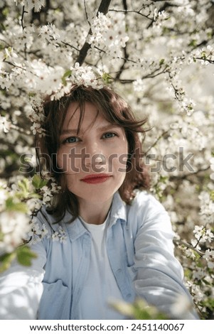 Portrait of a beautiful young woman in a blue shirt in cherry blossom tree. Gentle spring girl, natural, romantic. White flowers. Selfie, looking at camera. Vertical photo