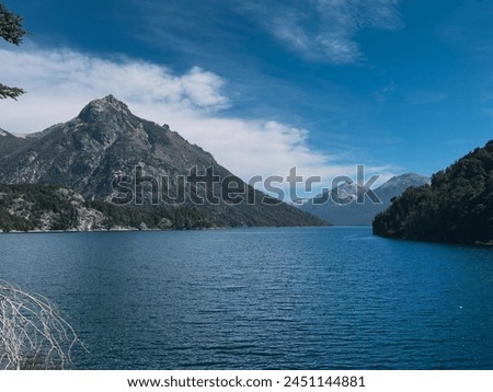 A beautiful lake with mountains in the background. The water is calm and clear. The sky is blue and there are some clouds. The mountains are covered in trees