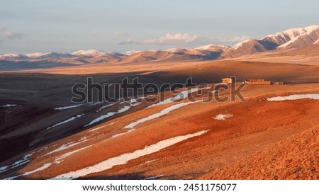 Bright sunny panoramic steppe landscape with a wooden barn. Wooden house on red dry grass, a mountain landscape of a wide prairie valley with a mountain range covered with snow. Perfect autumn image.