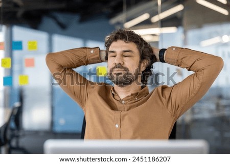 Calm caucasian guy sitting on chair with hands behind head and closed eyes in background of stylish office. Digital designer daydreaming and setting mind up for creative mood at beginning of workday.