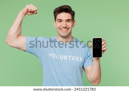 Young man wear blue t-shirt white title volunteer use blank screen mobile cell phone show muscles biceps isolated on plain green background. Voluntary free work assistance help charity grace concept