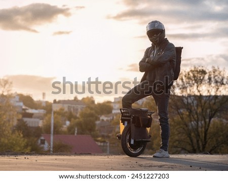 A young male rider in protective gear with a backpack on an electric unicycle against the background of the city at sunset. Driving around the city on a mono wheel Royalty-Free Stock Photo #2451227203