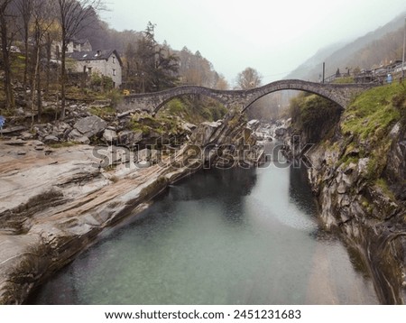 A photo of a stone bridge over a river in a mountainous area. The bridge is covered in greenery and the river below is flowing through a rocky gorge. The background consists of a foggy mountain