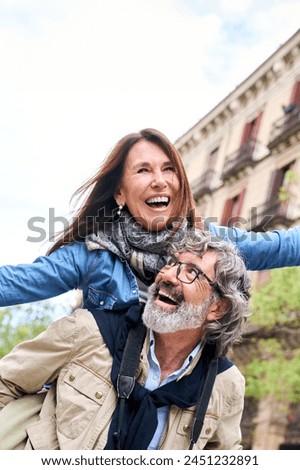 Vertical. Smiling elderly couple having fun together on vacation. Older man piggybacking mature woman enjoying leisure time visiting European city. Concept of excited people retirement relationships