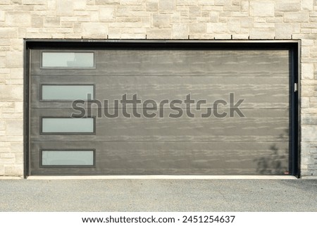 Dark grey double garage door on a residence with tan and beige textured brick. The modern style metal car garage door has four glass panel windows. The exterior automatic door rolls upward. Royalty-Free Stock Photo #2451254637