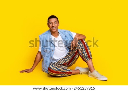 Full length photo of stylish handsome guy dressed jeans waistcoat print trousers sitting on floor isolated on yellow color background