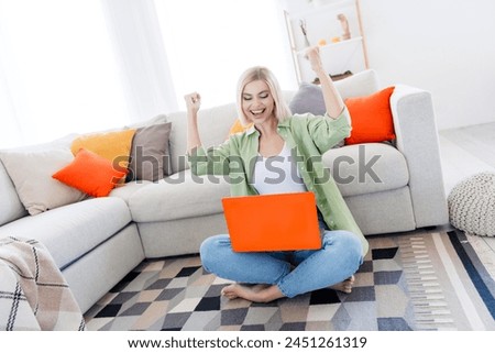 Photo of overjoyed cheerful girl sitting on floor indoors raising her fists chatting video call shouting yes success