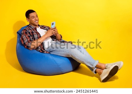Full size photo of good mood man dressed plaid shirt jeans trousers sit on pouf look at smartphone isolated on yellow color background