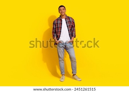 Full size photo of cool confident man dressed plaid shirt jeans trousers holding arms in pockets isolated on yellow color background