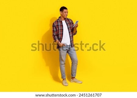 Full size photo of good mood man dressed plaid shirt jeans trousers look at smartphone read email isolated on yellow color background