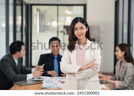Confident young businesswoman with arms crossed standing in office with colleagues discussing in background. Leadership and career concept