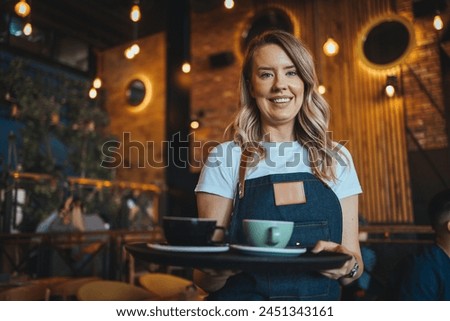 Nice woman waitress in apron, offering cup of delicious tasty coffee, stand smiling, friendly staff of restaurant.  Woman serving two cups of hot coffee to customer in cafeteria.  Royalty-Free Stock Photo #2451343161