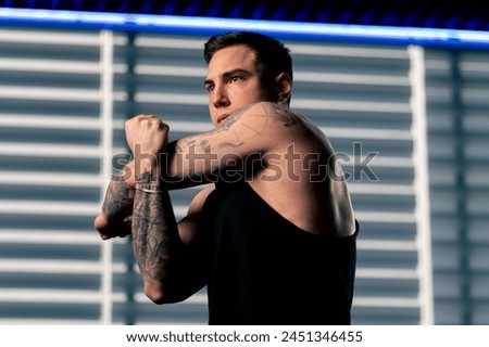 close up in the gym young handsome guy with tattoos in a black T-shirt is doing a warm-up in front of the exercise machine