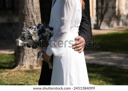 Newly married couple dressed as bride and groom, he in a black suit and she in a flowing white suit with a bouquet of flowers in her hand Royalty-Free Stock Photo #2451384099