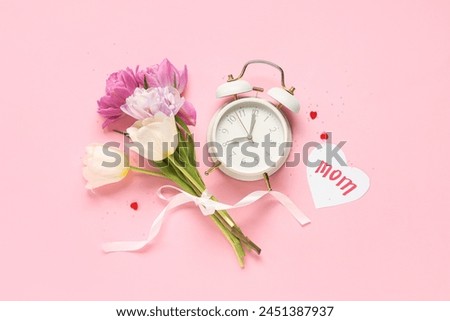 Card with word MOM, beautiful tulips and alarm clock on pink background. Mother's Day celebration