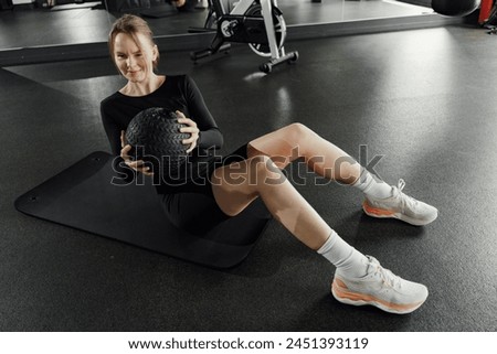 A woman with a wry smile performs a seated exercise on a gym mat, a medicine ball firmly grasped, showcasing active health.

 Royalty-Free Stock Photo #2451393119