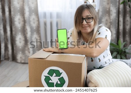 Smiling cute woman holds a phone with a chromakey screen in her hand over a box with a recycling sign. Clothes are packed in boxes with a recycling sign. Recycling old clothes.