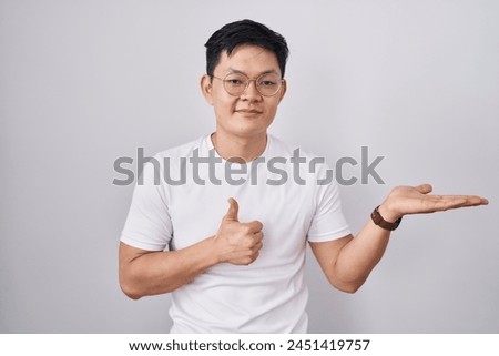 Young asian man standing over white background showing palm hand and doing ok gesture with thumbs up, smiling happy and cheerful 