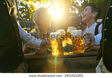 Friends in traditional German tracht toasting with bavarian beer mugs at sunset, happy expressions, in a outdoor setting in Beer garden or oktoberfest in Germany Royalty-Free Stock Photo #2451423303