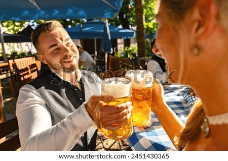 smiling man in traditional attire toasting with a woman in tracht at oktoberfest or beer garden in germany Royalty-Free Stock Photo #2451430365