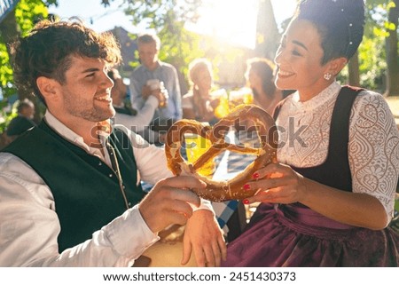 Couple in traditional German tracht holding a pretzel and smiling to each other at oktoberfest or beer garden in bavaria Royalty-Free Stock Photo #2451430373