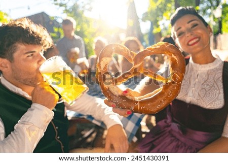 man drinking beer and a woman holding a giant pretzel, both in traditional German tracht, enjoying a sunny day at beer garden or oktoberfest Royalty-Free Stock Photo #2451430391