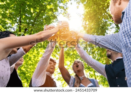 In Beer garden in Bavaria, Germany - friends in Tracht, Dindl and Lederhosen and Dirndl standing toasting beer mugs Royalty-Free Stock Photo #2451433709