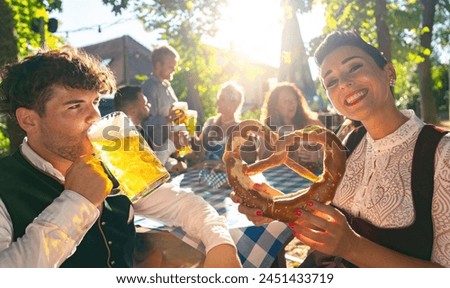 jovial woman and man in traditional German tracht with beer and a pretzel at a beer garden or oktoberfest in bavaria Royalty-Free Stock Photo #2451433719