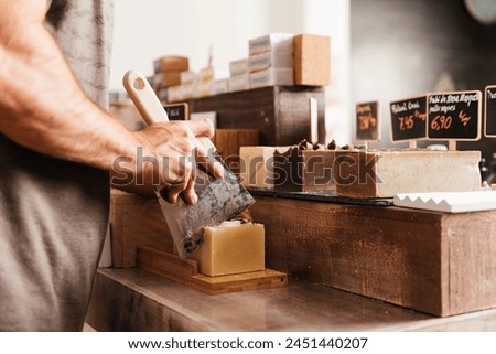 Close-up of a craftsperson slicing a homemade soap bar into pieces with a cutting tool on a wooden board. Text:"Mosqueta rose soap" Royalty-Free Stock Photo #2451440207