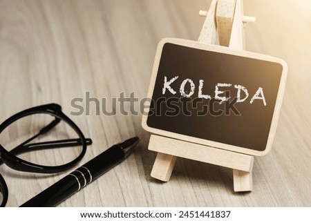 Black writing board on a wooden frame with the inscription "Kolęda", next to black glasses and a pen (selective focus) translation: carol